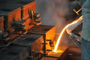 Molten metal is being poured into a mold, surrounded by steam and industrial equipment in a foundry setting.